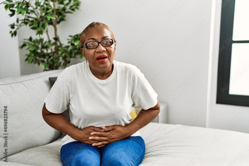 Mature hispanic woman sitting on the sofa at home with hand on stomach because indigestion, painful illness feeling unwell. ache concept. Mature hispanic woman sitting on the sofa at home with hand on stomach because indigestion, painful illness feeling unwell. ache concept.