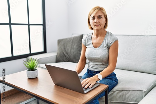 Middle age blonde woman using laptop at home relaxed with serious expression on face. simple and natural looking at the camera.