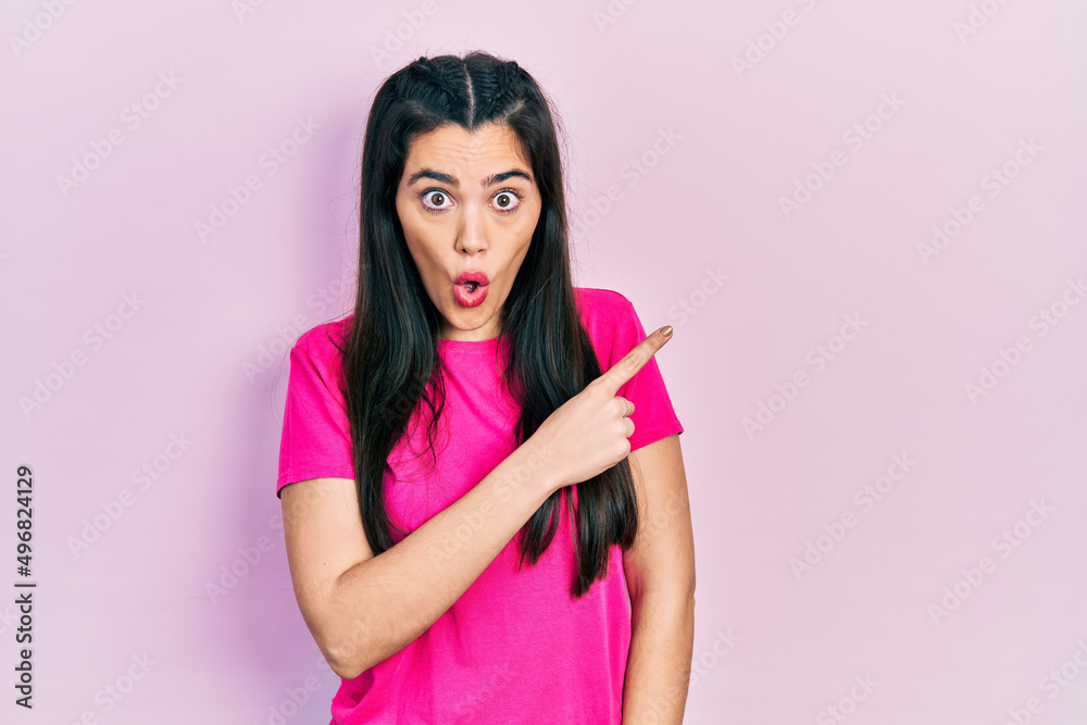 Young hispanic girl wearing casual pink t shirt surprised pointing with finger to the side, open mouth amazed expression. Young hispanic girl wearing casual pink t shirt surprised pointing with finger to the side, open mouth amazed expression.