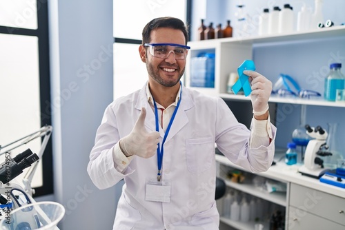 Young hispanic man with beard working at scientist laboratory holding blue ribbon smiling happy and positive  thumb up doing excellent and approval sign