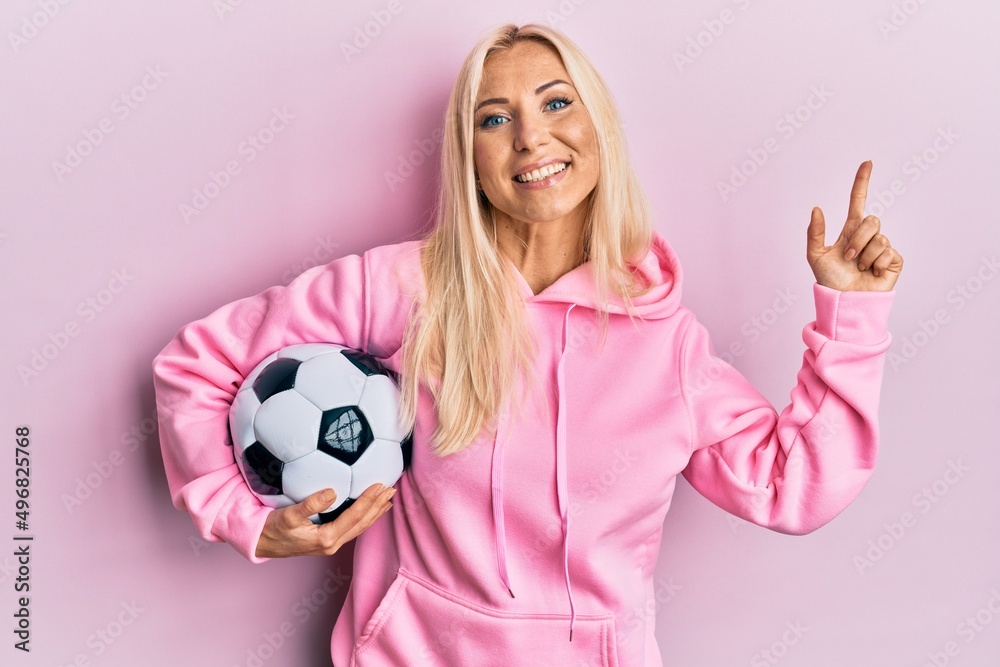 Young blonde woman holding soccer ball smiling happy pointing with hand and finger to the side Young blonde woman holding soccer ball smiling happy pointing with hand and finger to the side