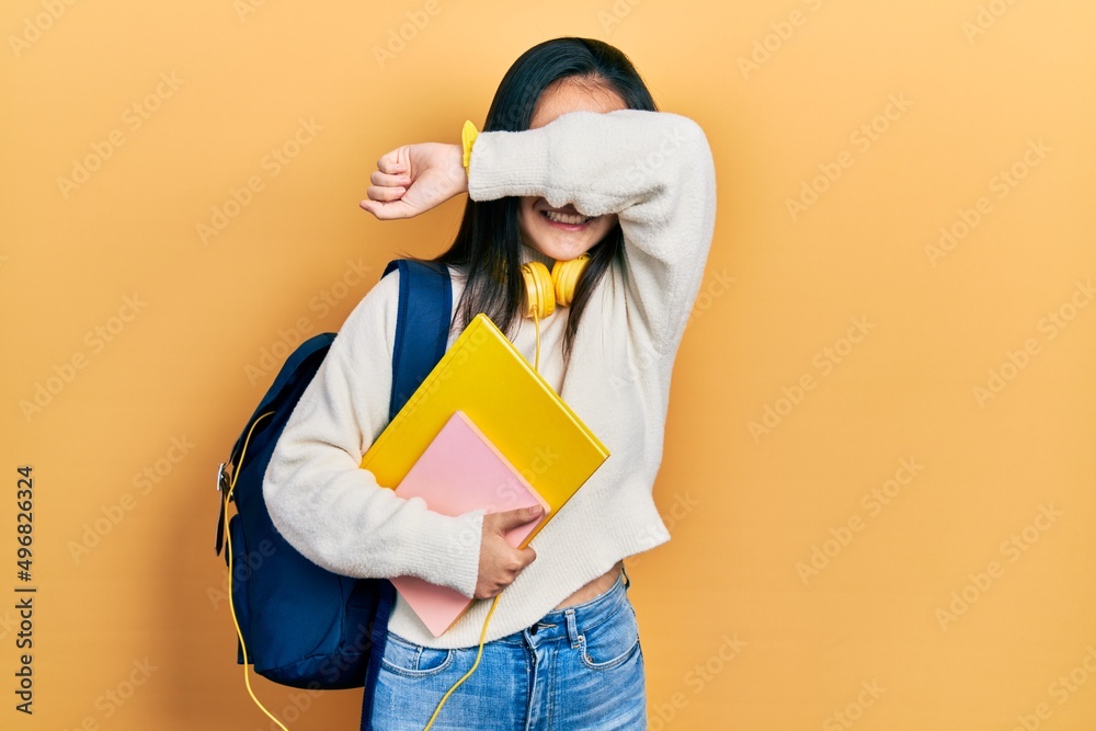 Young chinese girl holding student backpack and books smiling cheerful playing peek a boo with hands showing face. surprised and exited