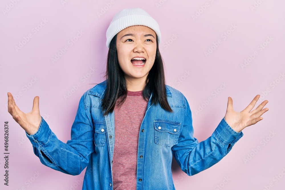 Young chinese girl wearing cute wool cap crazy and mad shouting and yelling with aggressive expression and arms raised. frustration concept. Young chinese girl wearing cute wool cap crazy and mad shouting and yelling with aggressive expression and arms raised. frustration concept.