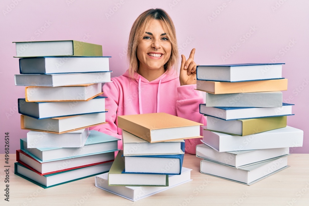 Young caucasian woman sitting on the table with books with a big smile on face, pointing with hand finger to the side looking at the camera. Young caucasian woman sitting on the table with books with a big smile on face, pointing with hand finger to the side looking at the camera.