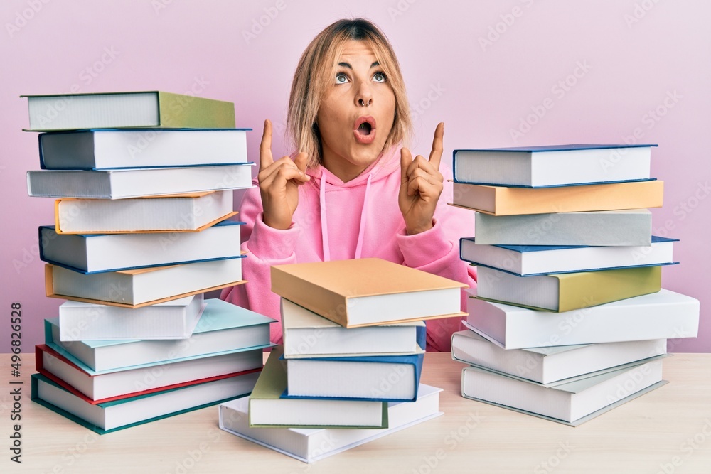 Young caucasian woman sitting on the table with books amazed and surprised looking up and pointing with fingers and raised arms. Young caucasian woman sitting on the table with books amazed and surprised looking up and pointing with fingers and raised arms.