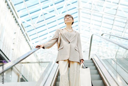Clean low angle of smart young woman in shopping mall going down escalator against glass ceiling, copy space