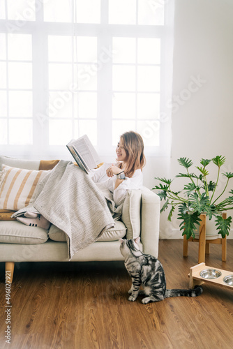 Young girl relaxing on chair playing with cute tabby cat while reading book in cabin near the window warm, cozy, domestic atmosphere.