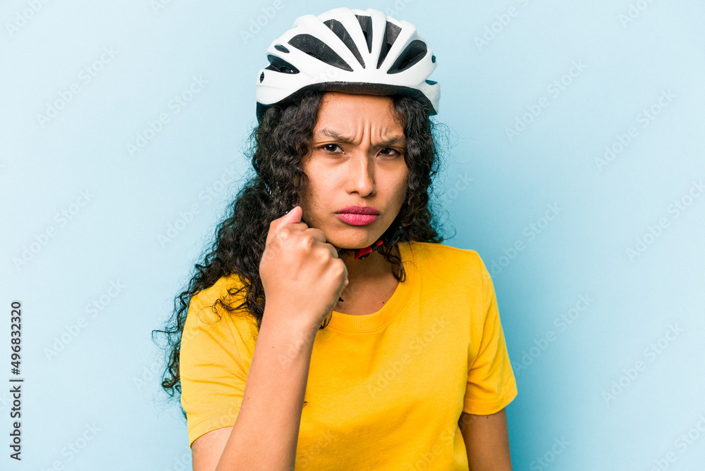 Young hispanic woman wearing a helmet bike isolated on blue background showing fist to camera, aggressive facial expression. Young hispanic woman wearing a helmet bike isolated on blue background showing fist to camera, aggressive facial expression.