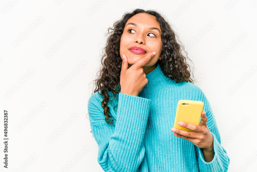 Young hispanic woman holding mobile phone isolated on white background looking sideways with doubtful and skeptical expression. Young hispanic woman holding mobile phone isolated on white background looking sideways with doubtful and skeptical expression.