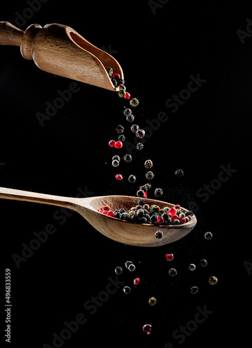 Fotografie Red and black peppercorns falling down into the wooden spoon at black background