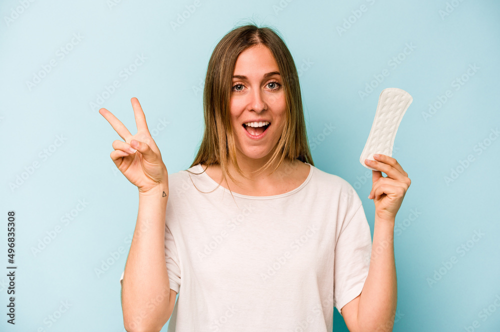 Young caucasian woman holding sanitary napkin isolated on blue background joyful and carefree showing a peace symbol with fingers. Young caucasian woman holding sanitary napkin isolated on blue background joyful and carefree showing a peace symbol with fingers.