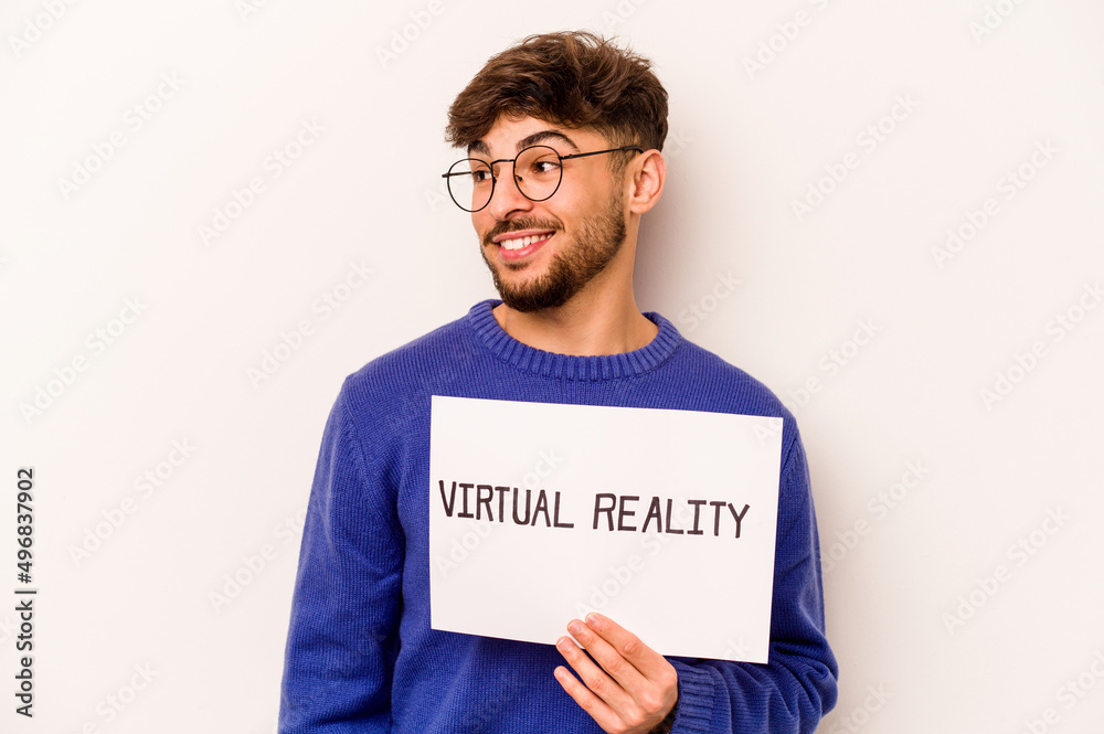 Young hispanic man holding a virtual reality placard isolated on white background looks aside smiling, cheerful and pleasant. Young hispanic man holding a virtual reality placard isolated on white background looks aside smiling, cheerful and pleasant.