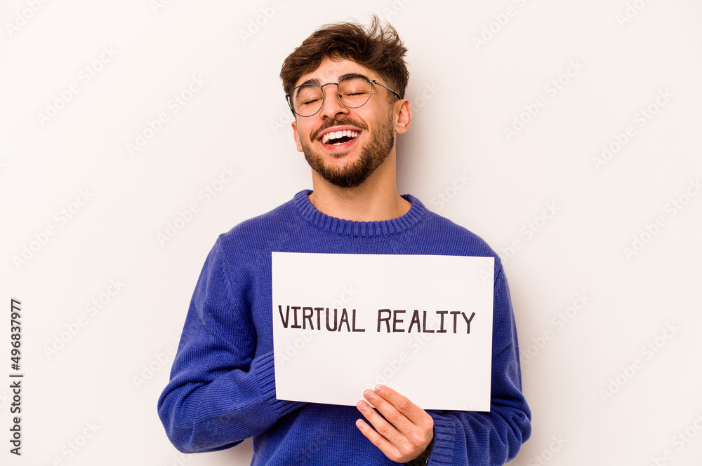 Young hispanic man holding a virtual reality placard isolated on white background laughs out loudly keeping hand on chest. Young hispanic man holding a virtual reality placard isolated on white background laughs out loudly keeping hand on chest.