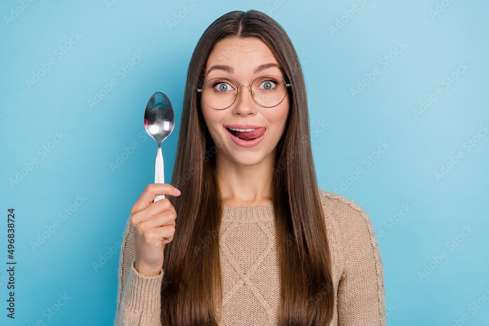 Photo of excited funny lady hold spoon protrude tongue lick lip wear specs beige sweater isolated blue color background Photo of excited funny lady hold spoon protrude tongue lick lip wear specs beige sweater isolated blue color background