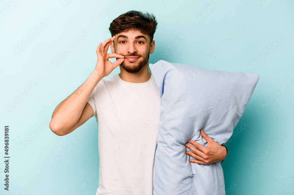 Young hispanic man wearing a pajama holding pillow isolated on blue background with fingers on lips keeping a secret.