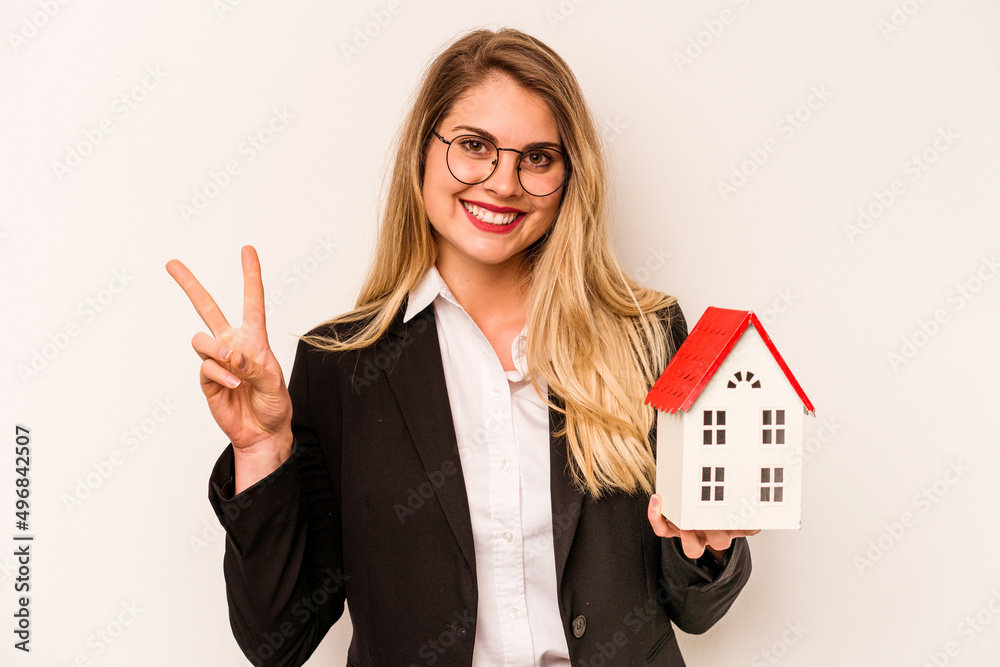 Young business caucasian woman holding a toy house isolated on white background joyful and carefree showing a peace symbol with fingers. Young business caucasian woman holding a toy house isolated on white background joyful and carefree showing a peace symbol with fingers.