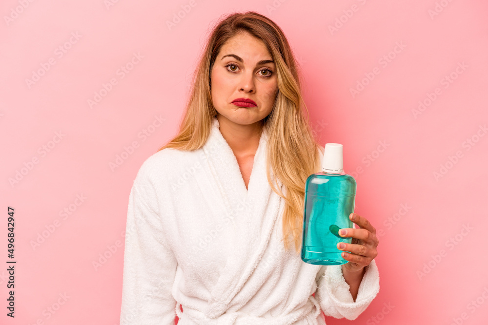 Young caucasian woman holding mouthwash wearing bathrobe isolated on pink background shrugs shoulders and open eyes confused. Young caucasian woman holding mouthwash wearing bathrobe isolated on pink background shrugs shoulders and open eyes confused.