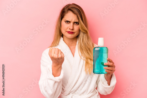 Young caucasian woman holding mouthwash wearing bathrobe isolated on pink background showing fist to camera, aggressive facial expression.