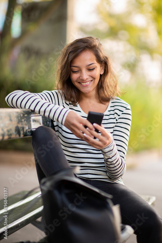 smiling young woman sitting on park bench looking at mobile phone © mimagephotos