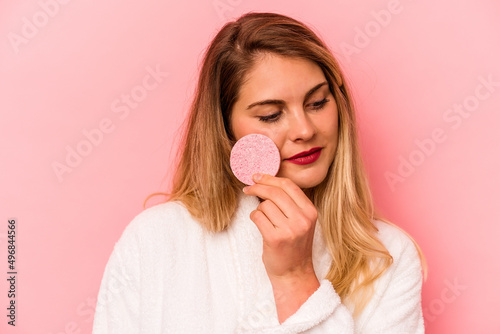 Young caucasian woman holding facial sponge isolated on pink background