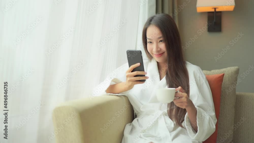 Beautiful asian woman in bathrobe on bright morning checking messages on mobile phone and holding cup of coffee Beautiful asian woman in bathrobe on bright morning checking messages on mobile phone and holding cup of coffee