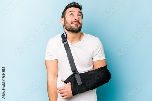 Young caucasian man with broken hand isolated on blue background dreaming of achieving goals and purposes