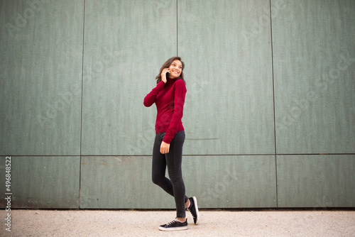 Full body young woman walking and talking with cellphone by wall