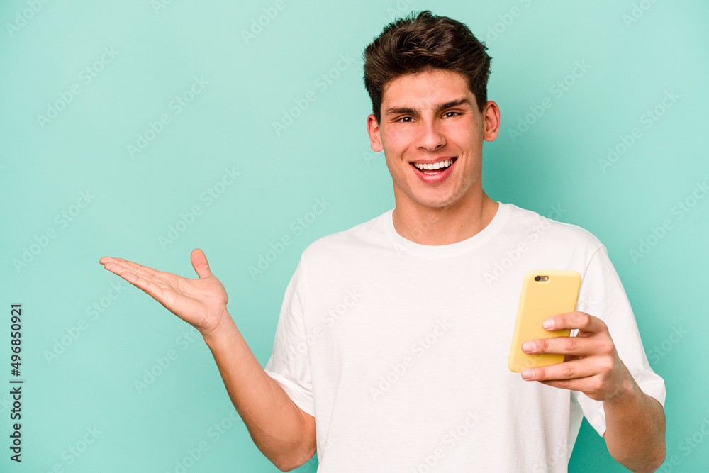 Young caucasian man holding mobile phone isolated on blue background showing a copy space on a palm and holding another hand on waist. Young caucasian man holding mobile phone isolated on blue background showing a copy space on a palm and holding another hand on waist.