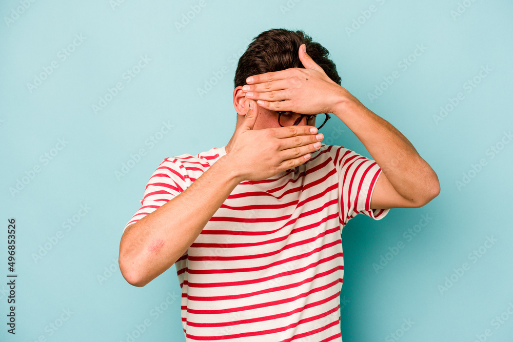 Young caucasian man isolated on blue background blink at the camera through fingers, embarrassed covering face. Young caucasian man isolated on blue background blink at the camera through fingers, embarrassed covering face.