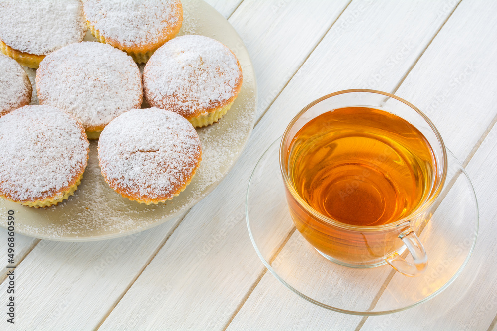 Freshly baked appetizing cupcakes sprinkled with powdered sugar on a plate and a cup of tea in a transparent cup on a white wooden table Freshly baked appetizing cupcakes sprinkled with powdered sugar on a plate and a cup of tea in a transparent cup on a white wooden table