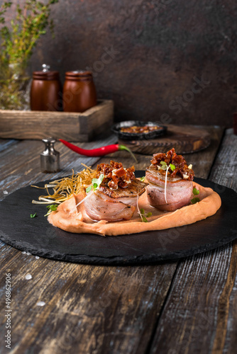 On a serving board, mashed sweet potatoes with bacon rolls with sun-dried tomatoes, microgreens and onion chips. On a wooden table is a box with spices and herbs, a glass of cognac. Restaurant menu