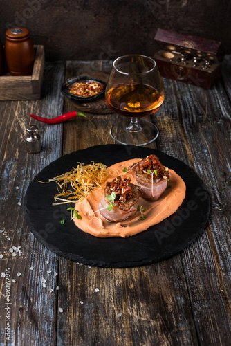 On a serving board, mashed sweet potatoes with bacon rolls with sun-dried tomatoes, microgreens and onion chips. On a wooden table is a box with spices and herbs, a glass of cognac. Restaurant menu