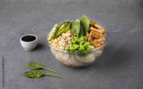 Poke bowl with rice, tofu, edamame beans, cucumber, chukka, spinach and peanuts on a gray textured background. Next to soy sauce and spinach leaves. Copy space. Close up. photo