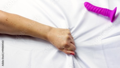 In a bed a woman grabs the sheets with her hand, next to it there is a pink dildo, concept of sexual pleasure