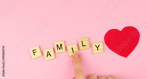 The word family from wooden letters and a red heart on a pink background. Female hand collects the word family and wooden letters