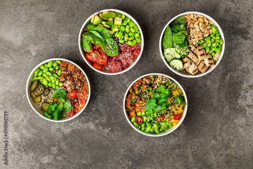 4 poke bowls with tofu, salmon, chicken, tuna, cucumbers, salad leafs, edamame beans, spinach and peanuts on a gray textured background. Top view.