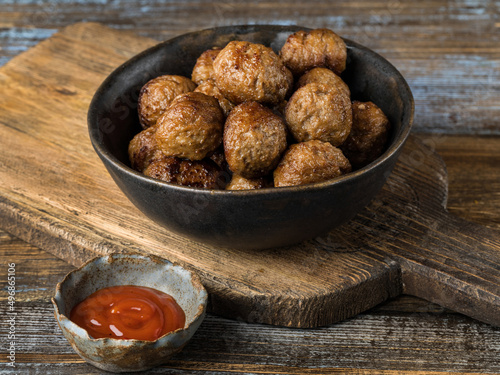 Fried beef meatballs in a dark ceramic bowl. Wooden brown background. Next to a cup of ketchup. Close up.