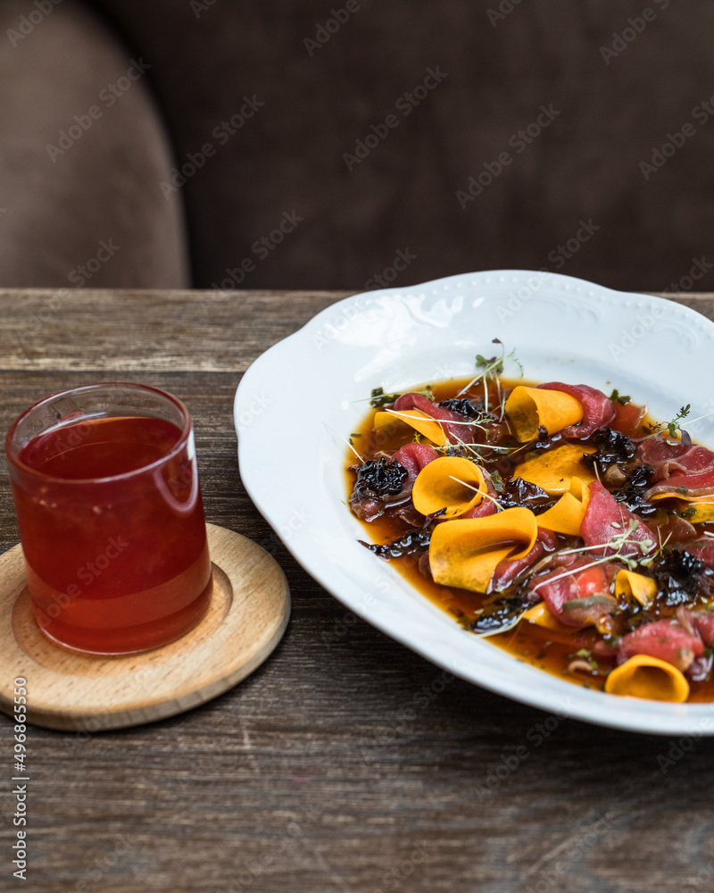 Oval plate with beef carpaccio with prunes, wine, sweet potatoes and micro greenery. On a wooden table in a restaurant, a transparent mug with red tea. Oval plate with beef carpaccio with prunes, wine, sweet potatoes and micro greenery. On a wooden table in a restaurant, a transparent mug with red tea.