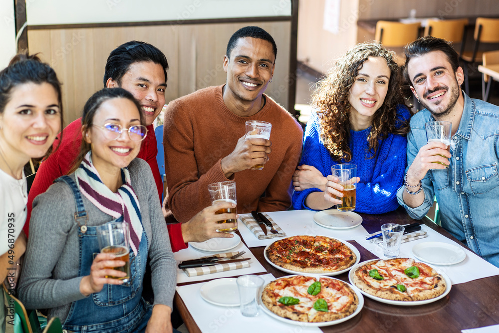 Smiling friends looking at camera while eating pizza at modern pizzeria restaurant - Friendship concept with multi ethnic people enjoying time together having fun at pizzeria with pizza and beer pints
