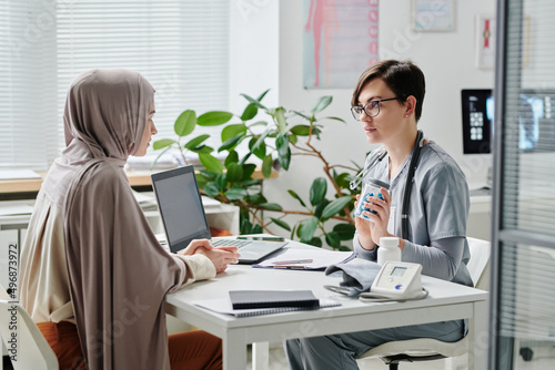 Young female doctor in blue uniform recommending new effective medicaments to Muslim patient in hijab in medical office