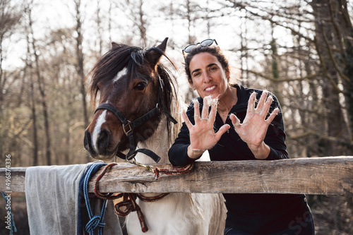 Beautiful smile woman standing next to her cute pony and showing hands full with horse hair in spring shedding season on countryside ranch. Horse molt shower.