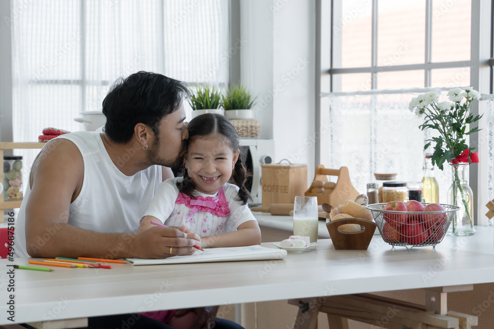 Happy Asian young father with beard kissing head of little cute daughter while drawing with colored pencil together at home with glass of milk, marshmallows, breads and apples on kitchen table Happy Asian young father with beard kissing head of little cute daughter while drawing with colored pencil together at home with glass of milk, marshmallows, breads and apples on kitchen table