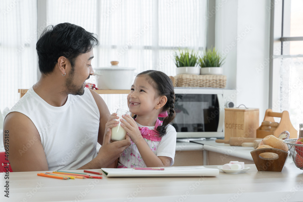 Happy Asian young father with beard holding feeding glass of milk for little cute daughter drinking while drawing with colored pencil together at home with marshmallows and breads on kitchen table Happy Asian young father with beard holding feeding glass of milk for little cute daughter drinking while drawing with colored pencil together at home with marshmallows and breads on kitchen table