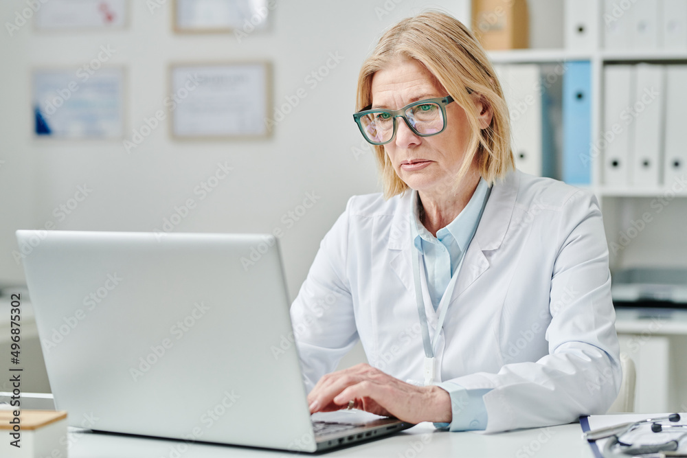 Serious mature blond female doctor in whitecoat and eyeglasses typing on laptop keyboard while sitting by desk in medical office Serious mature blond female doctor in whitecoat and eyeglasses typing on laptop keyboard while sitting by desk in medical office