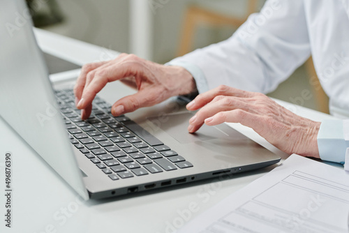 Hands of mature female clinician pressing buttons of laptop keyboard while sitting by desk and consulting patients online