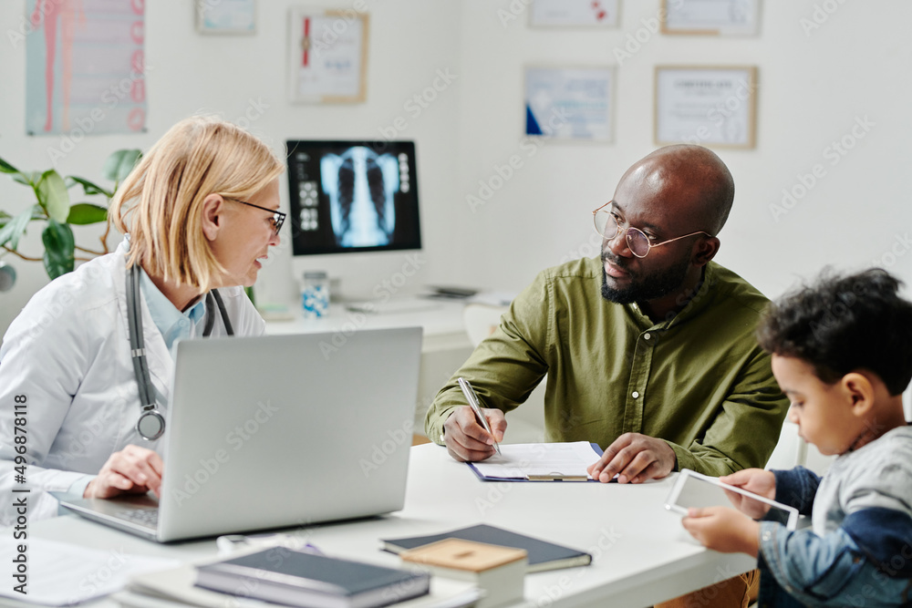 Experienced doctor with laptop consulting African American male patient filling in medical form while his cute little son using tablet Experienced doctor with laptop consulting African American male patient filling in medical form while his cute little son using tablet