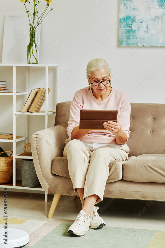 Vertical full length portrait of modern senior woman using tablet while sitting on sofa in white home interior