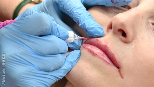 Skilled cosmetologist applying permanent makeup on lips in a beauty salon. Woman undergoing procedure of lip blushing with tattoo machine. Close-up
