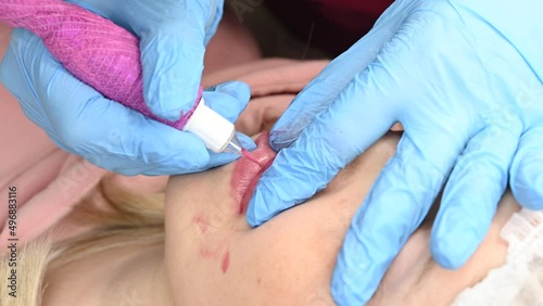Skilled cosmetologist applying permanent makeup on lips in a beauty salon. Woman undergoing procedure of lip blushing with tattoo machine. Close-up