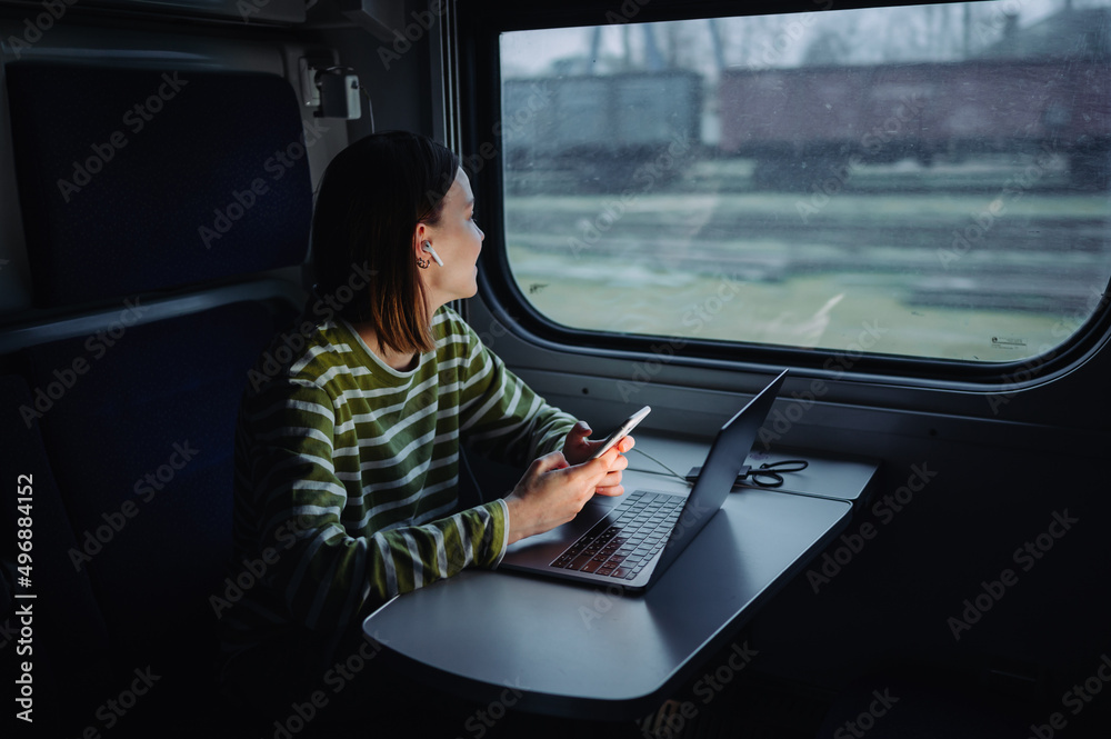 Positive woman in headphones sitting with a smartphone in her hands and using a laptop while traveling, looking out the window